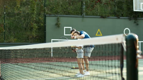 man teaching his little daughter how to hit ball with racket on a tennis court on a summer day