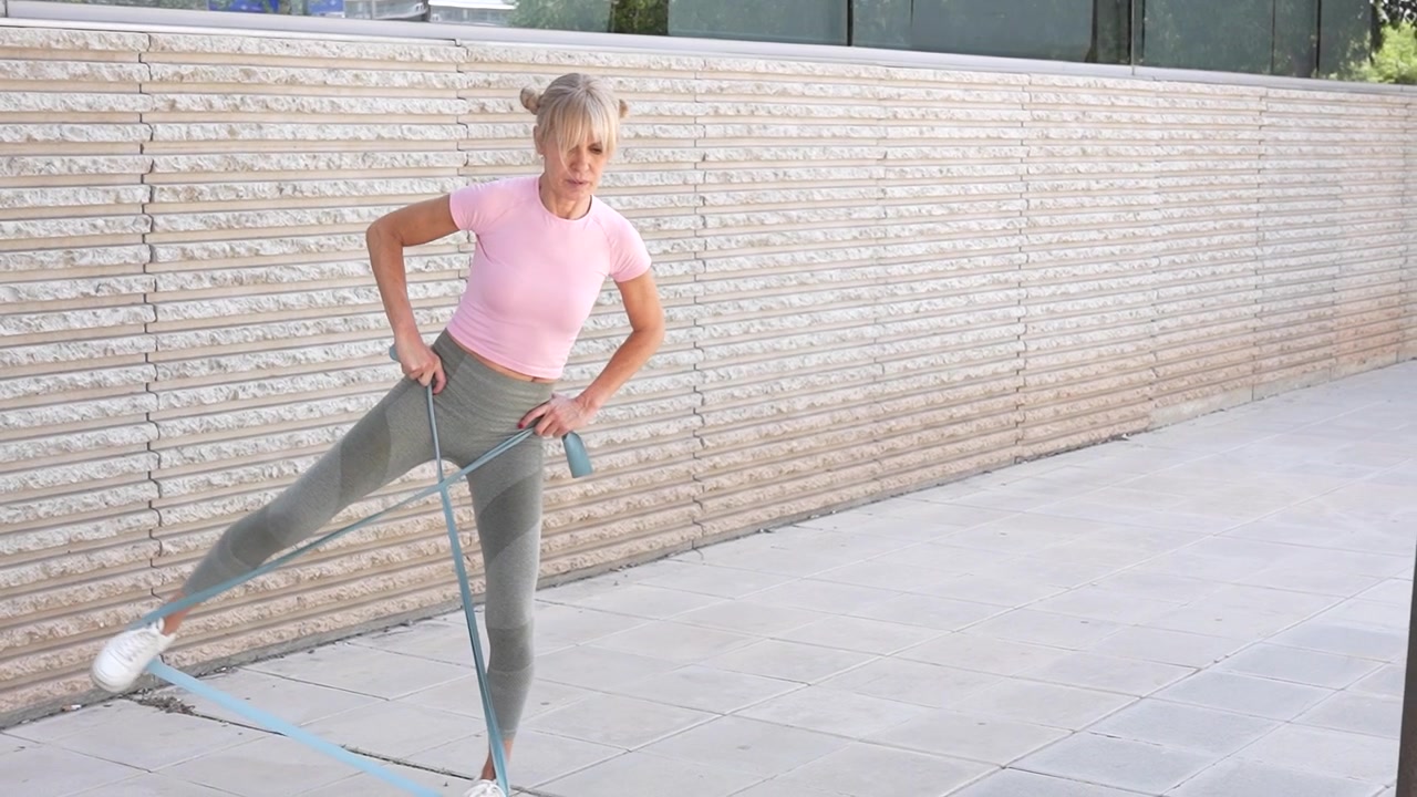 Woman exercises outdoors with resistance band dedicated to fitness in pink top and grey leggings