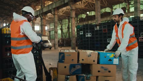 A-Black-man-in-a-white-uniform-together-with-his-colleague-a-man-with-a-beard-and-an-orange-vest-are-trying-to-put-a-wheelbarrow-with-sorted-waste-in-place-near-boxes-with-sorted-glass-waste-at-a-waste-recycling-plant