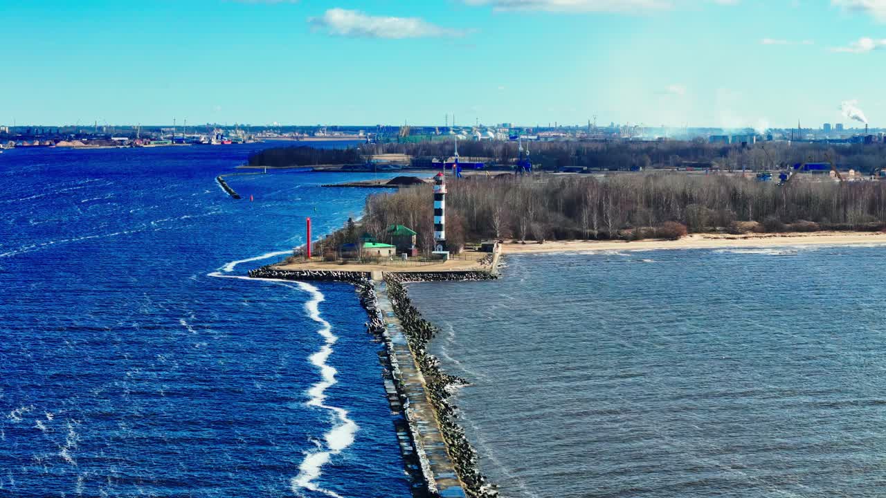 A striped lighthouse marks the entrance to a river where two bodies of water meet. A stone jetty cuts through the waves, with forest and beach in the background under a bright blue sky