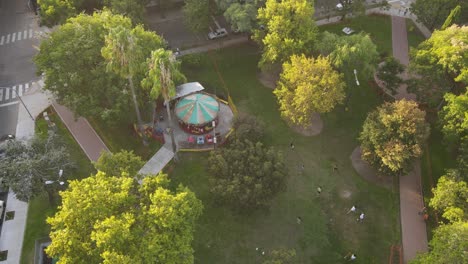 aerial top down of spinning carousel in tropical park at sunset time - people walking on path in buenos aires city