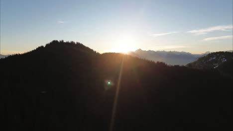 Drone-fly-above-dark-Mountain-Forest-and-reveals-Sunrise-behind-snow-covered-Mountain-Range