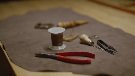 a set of leather crafting tools arranged on brown fabric, featuring a spool of waxed thread, an awl, snips, pliers, a beeswax block with needles, and metal hardware.