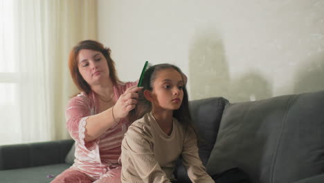 mother brushing daughter’s hair in cozy living room at home. quiet, intimate moment between parent and child, showing warmth, care, and connection. soft natural light coming through curtains