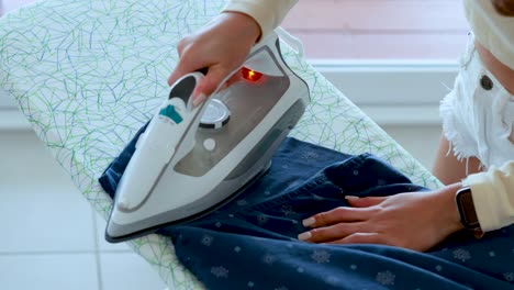 a woman uses a steam iron to press a blue shirt on an ironing board in a bright, naturally lit room, with smooth camera movement