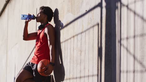 Fit-african-american-man-exercising-in-city-taking-a-break,-drinking-water,-holding-basketball