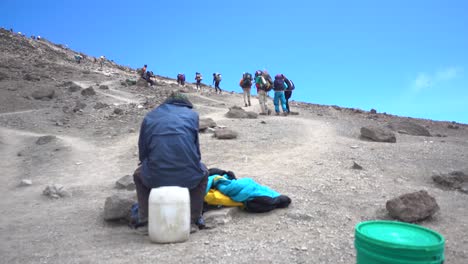 hombre sentado y descansando en la pista del monte kilimanjaro mientras los excursionistas caminan hacia arriba contra un cielo azul