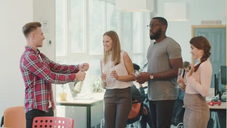 happy team drinking alcohol in workplace. young man opening bottle of champagne.