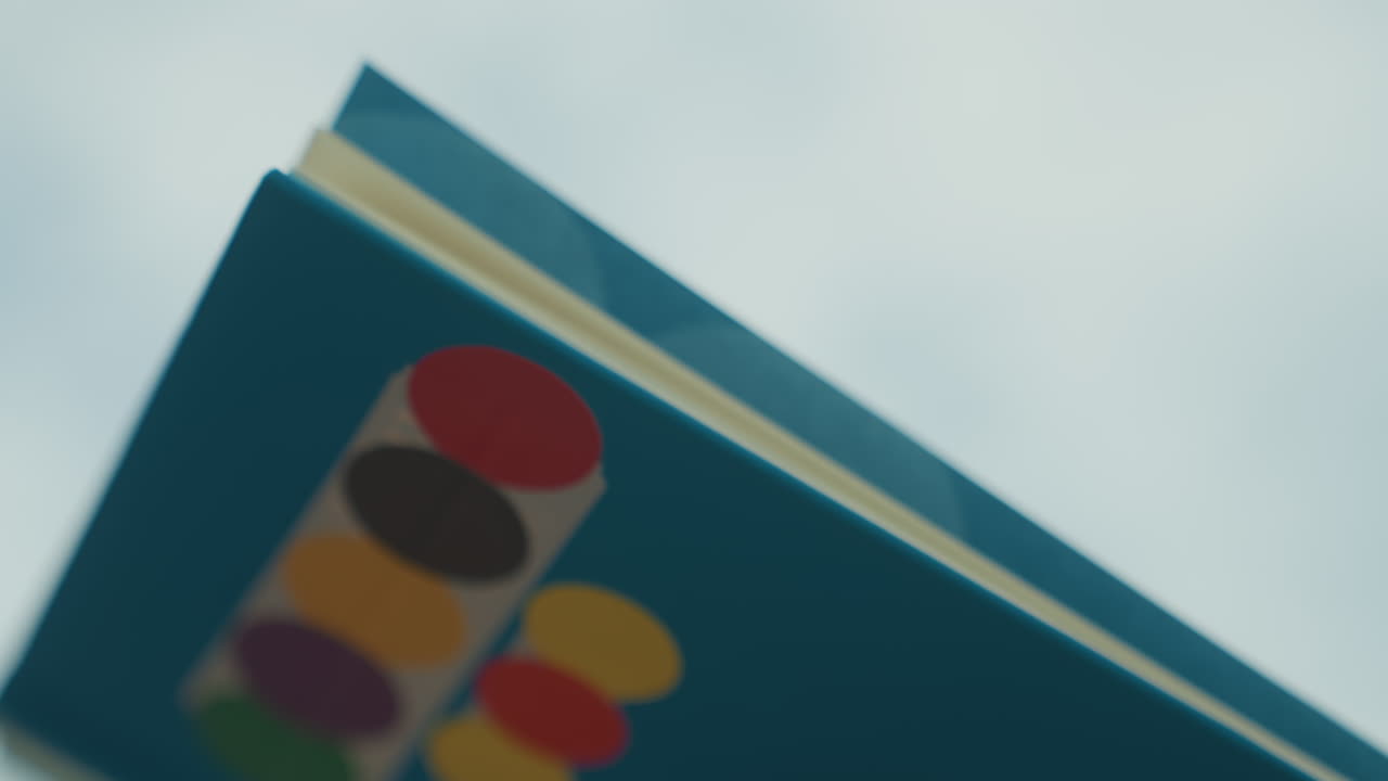 Blå boghjørne mod himlen, filmisk nærbillede, der viser et turkis paperback-omslag med flerfarvede klistermærkeprikker, skrå komposition, lav dybdeskarphed, blød, skyet bokeh-baggrund, synligt papir.