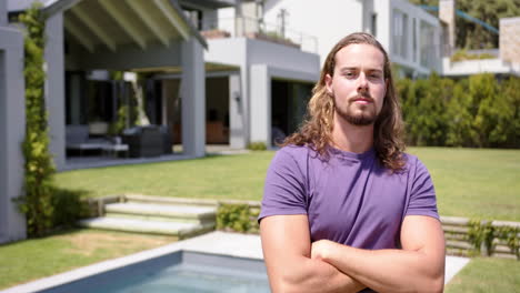 caucasian young man with arms crossed stands by pool outside in garden, copy space