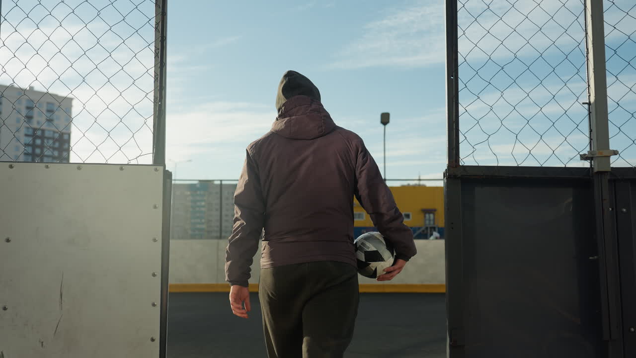 Back view of man walking away at an urban sports arena, carrying a soccer ball towards a gate, with cityscape and yellow storage containers in the background