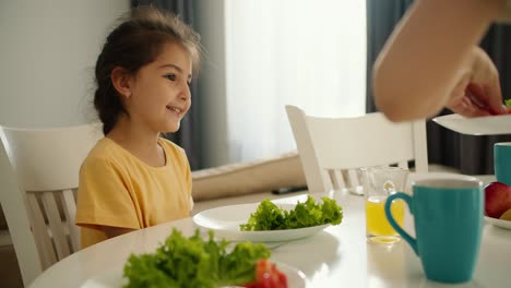 A-little-brunette-girl-in-a-yellow-dress-sits-on-a-chair-in-the-kitchen-and-her-mother-puts-green-herbs-and-vegetables-on-the-girl-s-plate-for-breakfast-at-the-table-in-the-morning