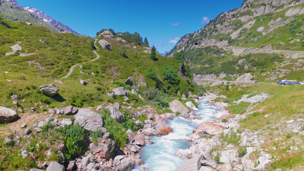 Natura selvaggia del Passo del Susten in Svizzera, ruscello di montagna che scorre attraverso una valle rocciosa e verde sotto un cielo azzurro limpido, con erba selvatica, massi e ripide colline in una soleggiata giornata estiva.