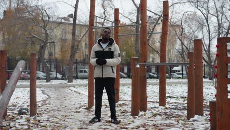 young adult performing neck twist outdoors in winter jacket, hands joined, standing amidst workout equipment in urban park, snow-covered ground, and bare trees in background