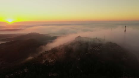 Drone-at-sunrise-flying-around-Monte-Toro-in-Menorca,-Spain-with-fog-rolling-over-the-mountains