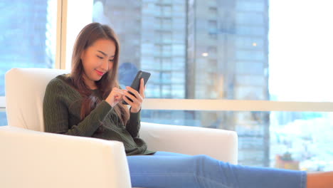 Una-Mujer-Joven-Se-Sienta-Cómodamente-En-Un-Sillón-Respaldado-Por-Modernos-Rascacielos-En-La-Ventana-Mientras-Toca,-Se-Desplaza-E-Interactúa-Con-Su-Teléfono-Inteligente