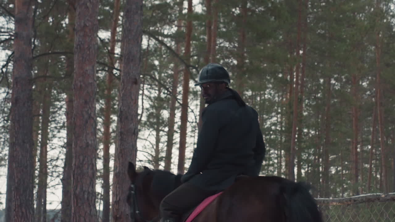 hombre negro montando a caballo en un pinar al atardecer, jinete con casco caminando por un sendero junto al lago, luz invernal y ambiente tranquilo, abrigo oscuro y silla de montar visibles, soledad reflectante al aire libre