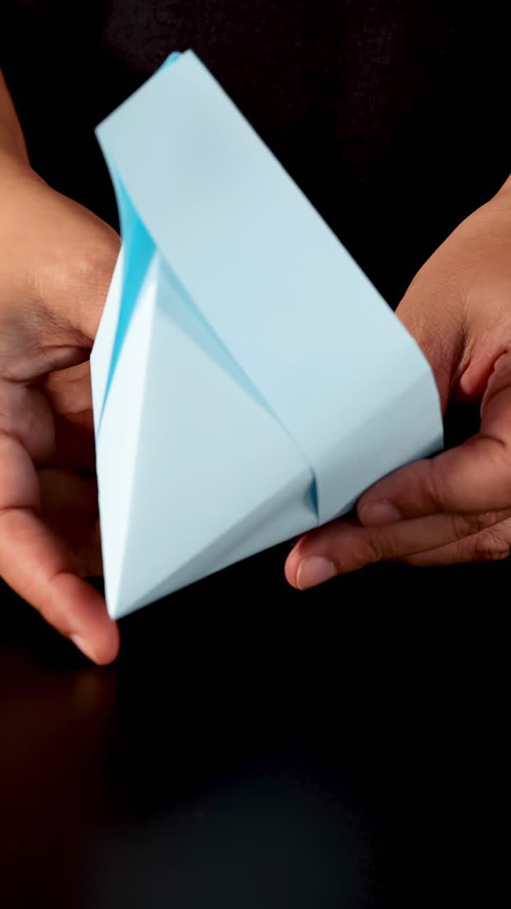 Mani piegano carta blu in una barca di origami su sfondo scuro, primo piano, illuminazione soffusa.