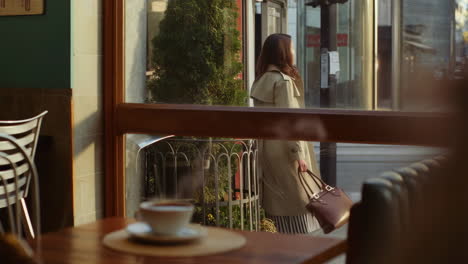 Woman in trench coat walking past a cafe window with coffee inside