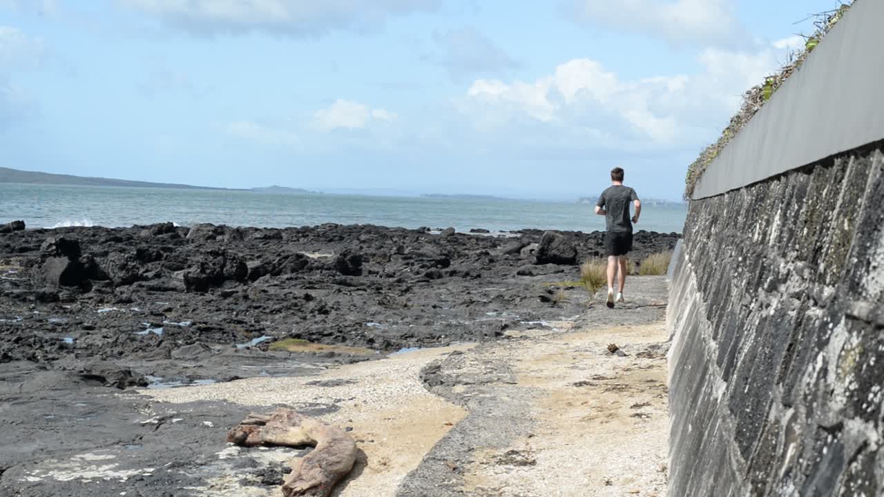 corredor masculino adulto desde atrás corriendo entre el malecón y las rocas volcánicas en la costa norte del pacífico de nueva zelanda