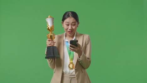 asian business woman in a suit with a gold medal and trophy looking at smartphone then saying wow on green screen background in the studio