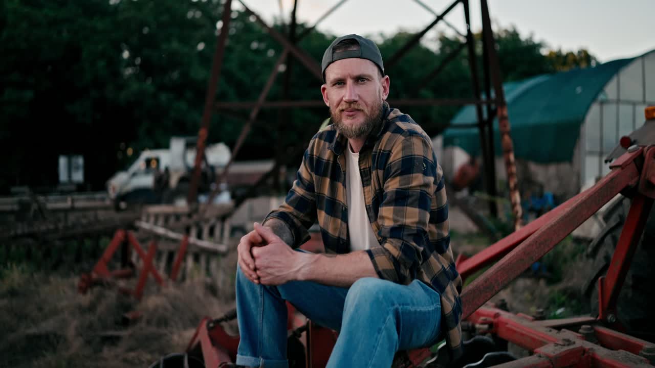 retrato de un feliz granjero en una gorra y camisa a cuadros que está sentado en una cosechadora combinada y posando cerca de un campo en una granja