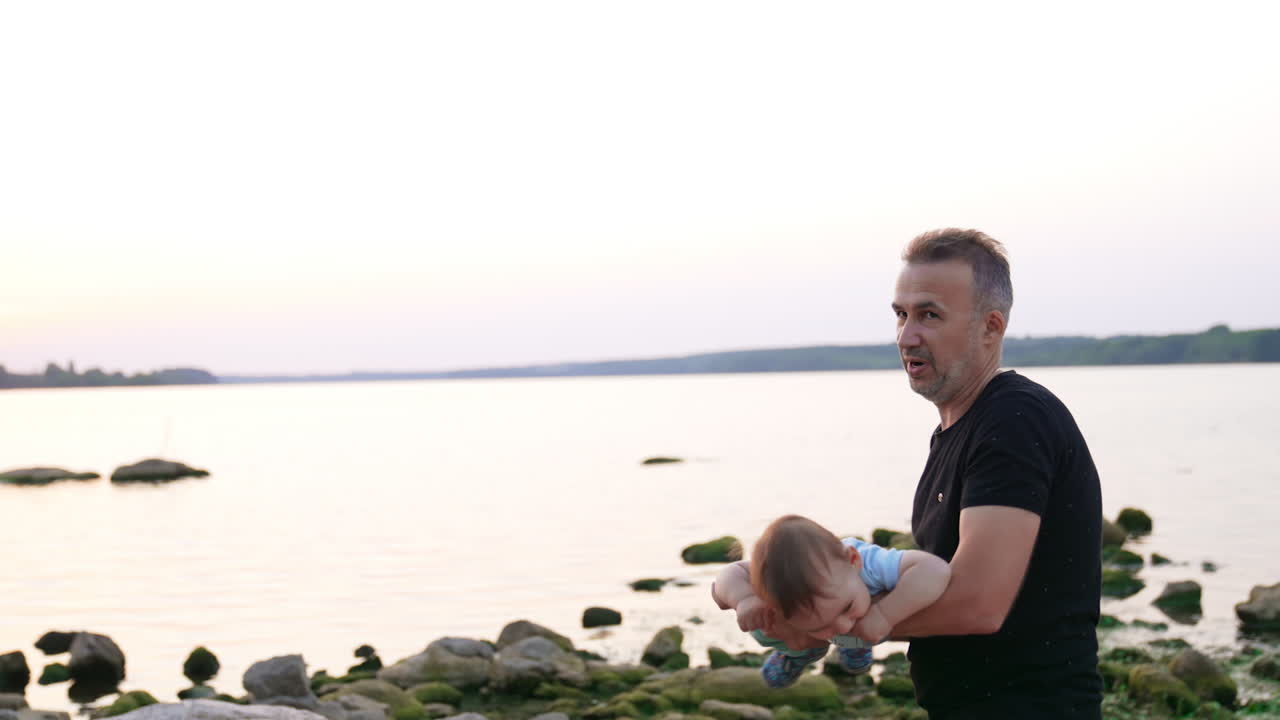 Sweet little kid flies lying on his dad's arms. Adorable child smiling holding his finger in mouth. Lake at backdrop.