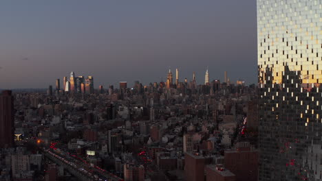 Fliegen-Sie-In-Der-Abenddämmerung-über-Die-Stadt,-Hohe-Beleuchtete-Wolkenkratzer-In-Der-Ferne.-Verschiebbare-Laibung-Eines-Glänzenden-Hochhauses,-Das-Den-Farbenprächtigen-Sonnenuntergangshimmel-Widerspiegelt.-Manhattan,-New-York-City,-Vereinigte-Staaten