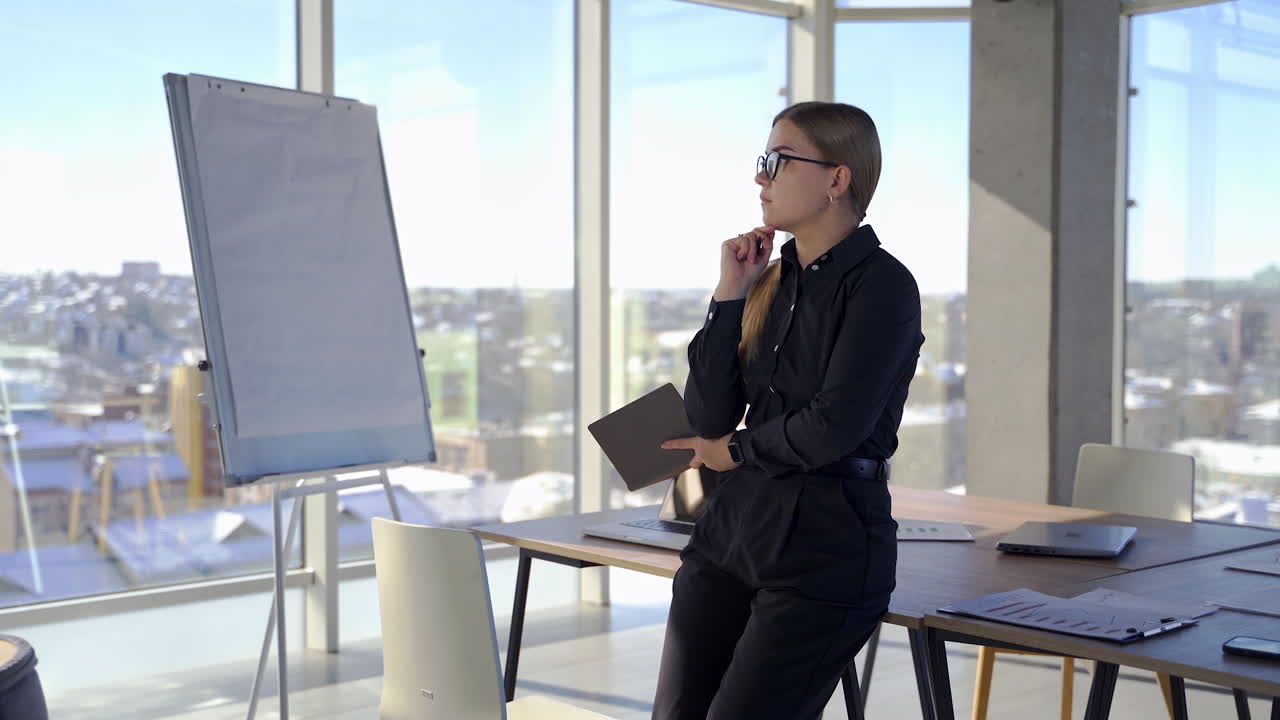 Pensive business woman in modern cabinet. Young pretty woman in black clothes and glasses holding a pocketbook and thinks about new business project indoors.