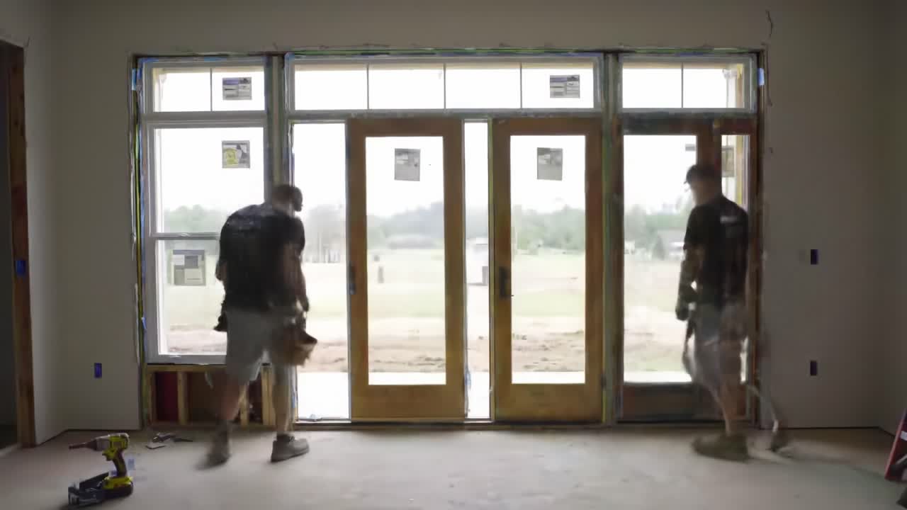 Two workers are engaged in renovation tasks within a home. One is installing components near a large window while another enters the space. Natural light brightens the area.