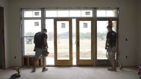 two workers are engaged in renovation tasks within a home. one is installing components near a large window while another enters the space. natural light brightens the area.