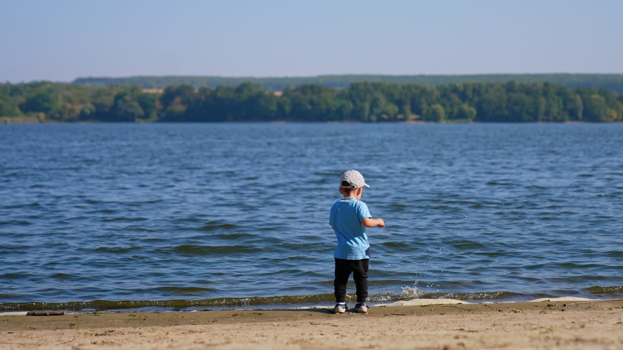 Baby boy comes up to river throwing some sand. Toddler having fun time on the river bank in summer.