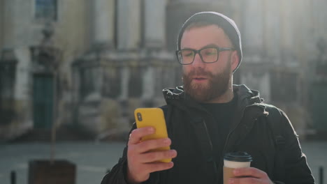 surprised man with beard and glasses looking at his smartphone outdoors while holding a coffee cup
