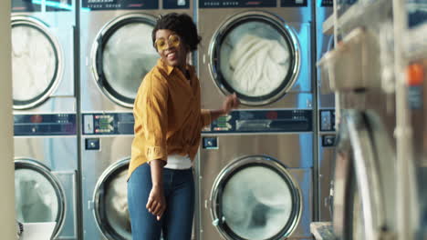 beautiful cheerful woman in stylish outfit having fun and dancing in laundry service room while machines washing on background