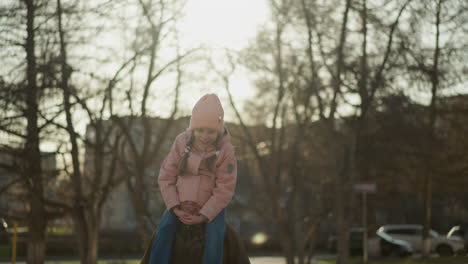 a little girl in a pink cap and jacket sits joyfully on her dad's neck, with half of his head snugly tucked in her jacket covering his eyes. the background features a sunlit park with bare trees