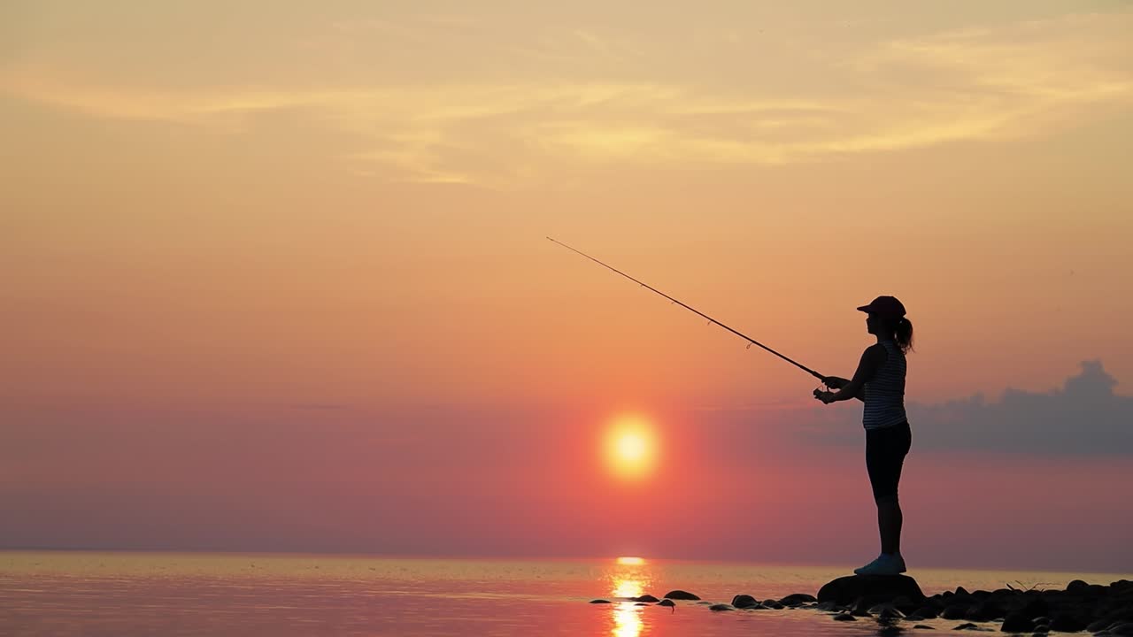 Wanita memancing dengan joran pancing berputar dengan latar belakang matahari terbenam.