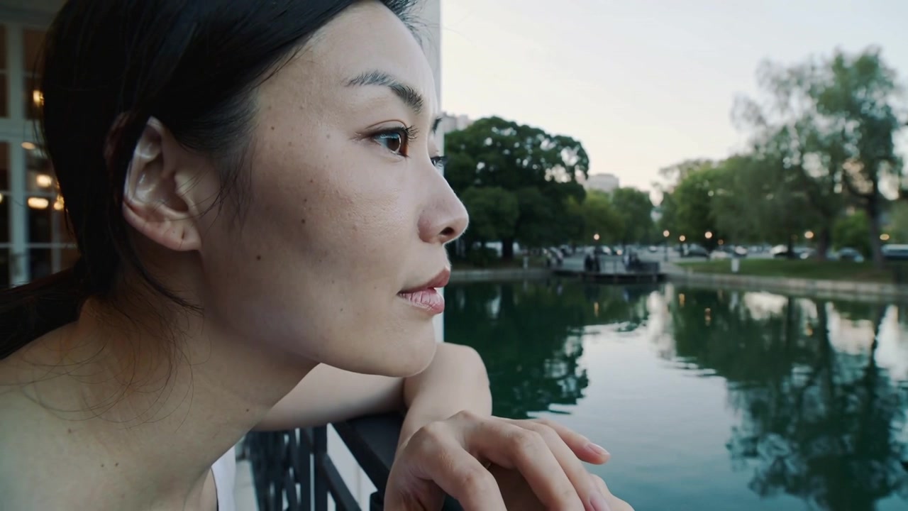 ノースリーブのトップスを着たアジア人女性が、金属製の手すりに寄りかかり、夕暮れが迫る中、池を眺めている。コピースペースあり。