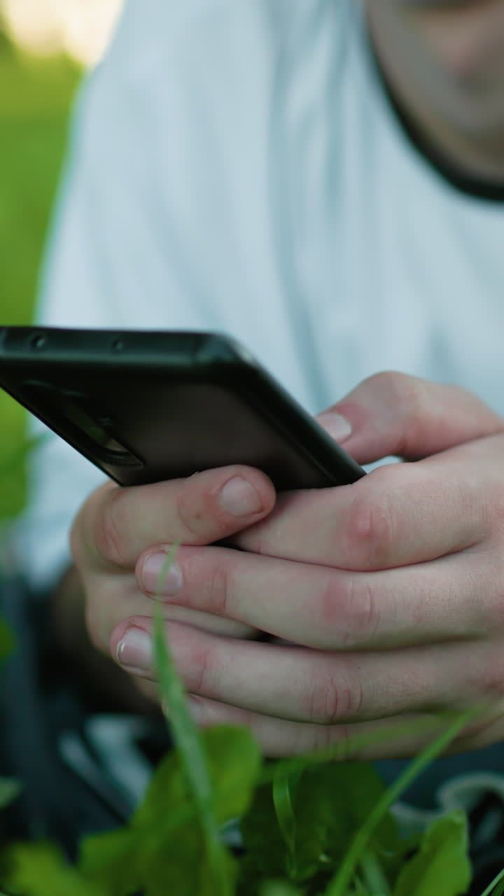 primer plano de un hombre tendido en un campo de hierba usando un teléfono inteligente con una mano tatuada visible, mostrando un momento pacífico al aire libre rodeado de exuberante vegetación