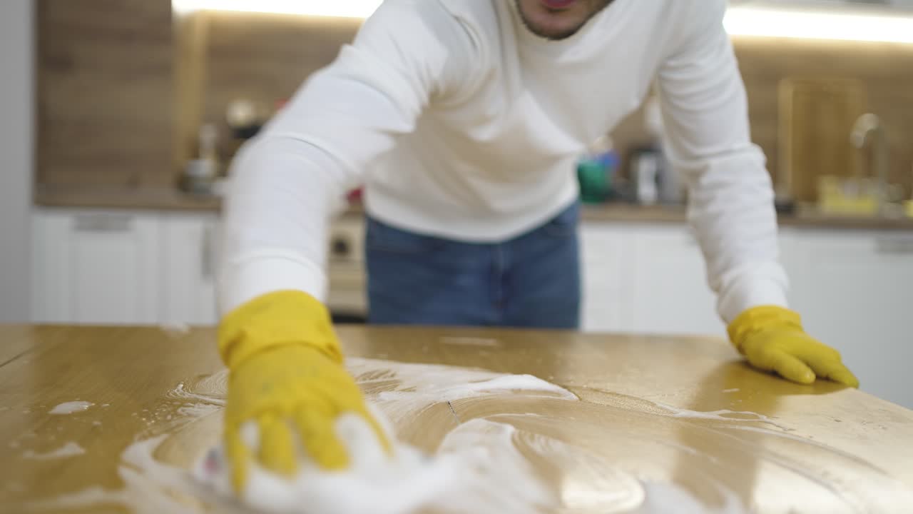 Hombre limpia una mesa con una esponja espumosa durante la limpieza de primavera, mostrando un cuidado exhaustivo del hogar, la limpieza de la cocina o sala de estar, y el mantenimiento doméstico diario en un entorno familiar.