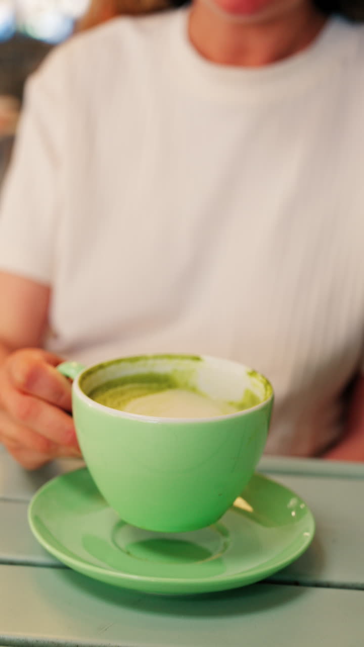 Close up of a blonde woman in a white T-shirt drinking a matcha latte out of a green cup at a terrace. Vertical