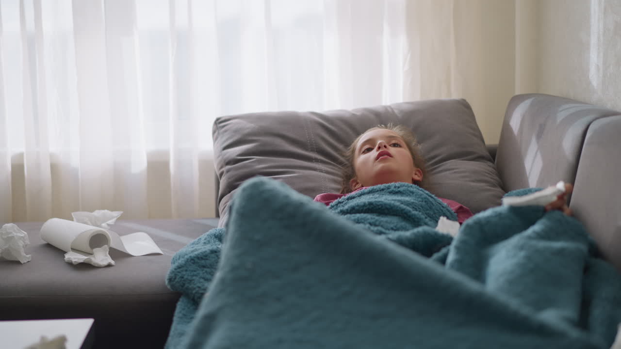 White girl on couch covered with soft blue blanket sneezing into tissue paper with tissues scattered around, wearing red bangle, showing signs of cold or flu, with tissues all over