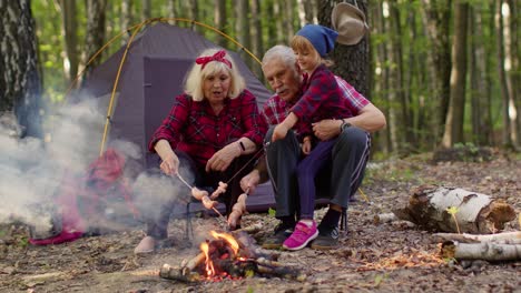 happy senior man woman with grandchild talking cooking frying sausages over campfire in evening wood