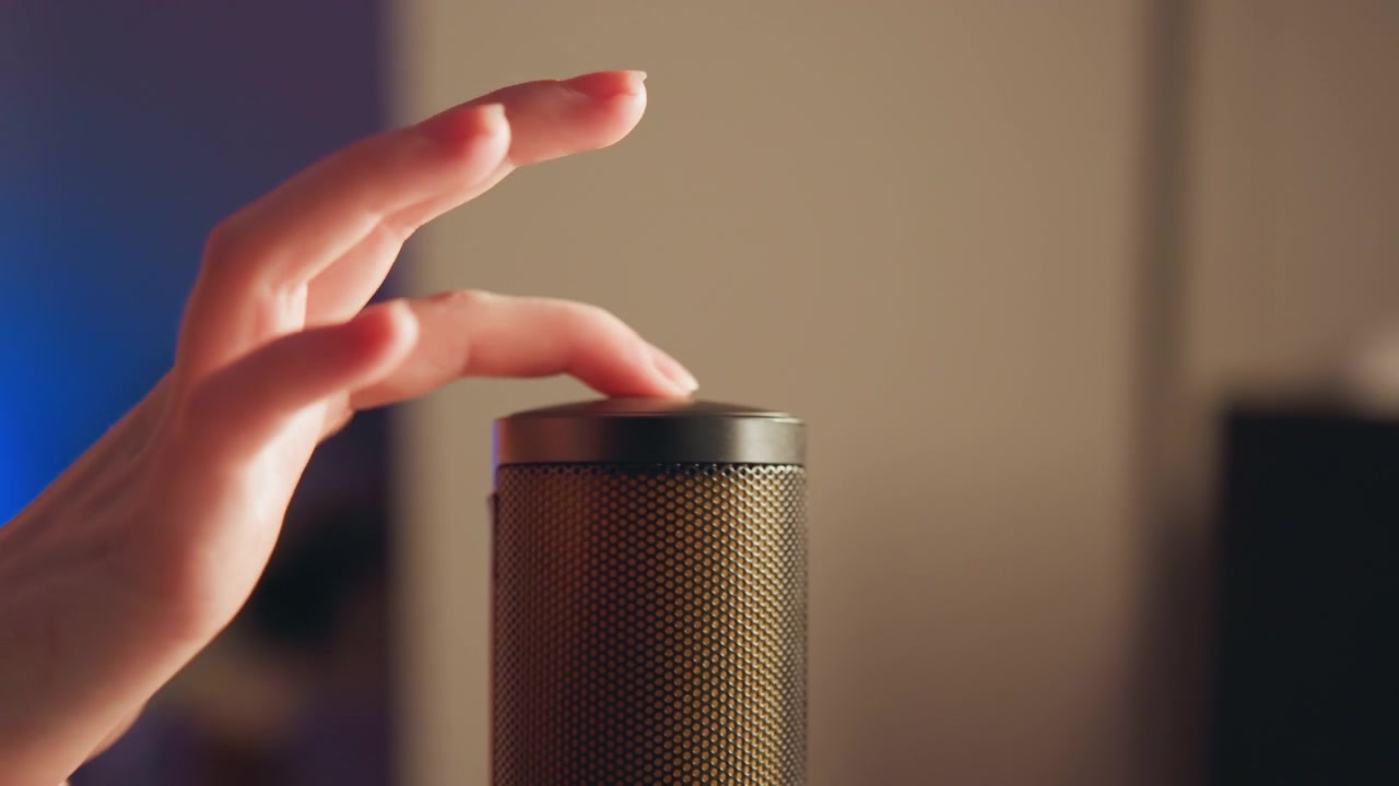 Microfone cilíndrico com grade de malha metálica e tampa fosca em primeiro plano, enquanto uma luz LED verde brilha na superfície e uma mão se estende da esquerda para ajustar ou tocar o dispositivo.