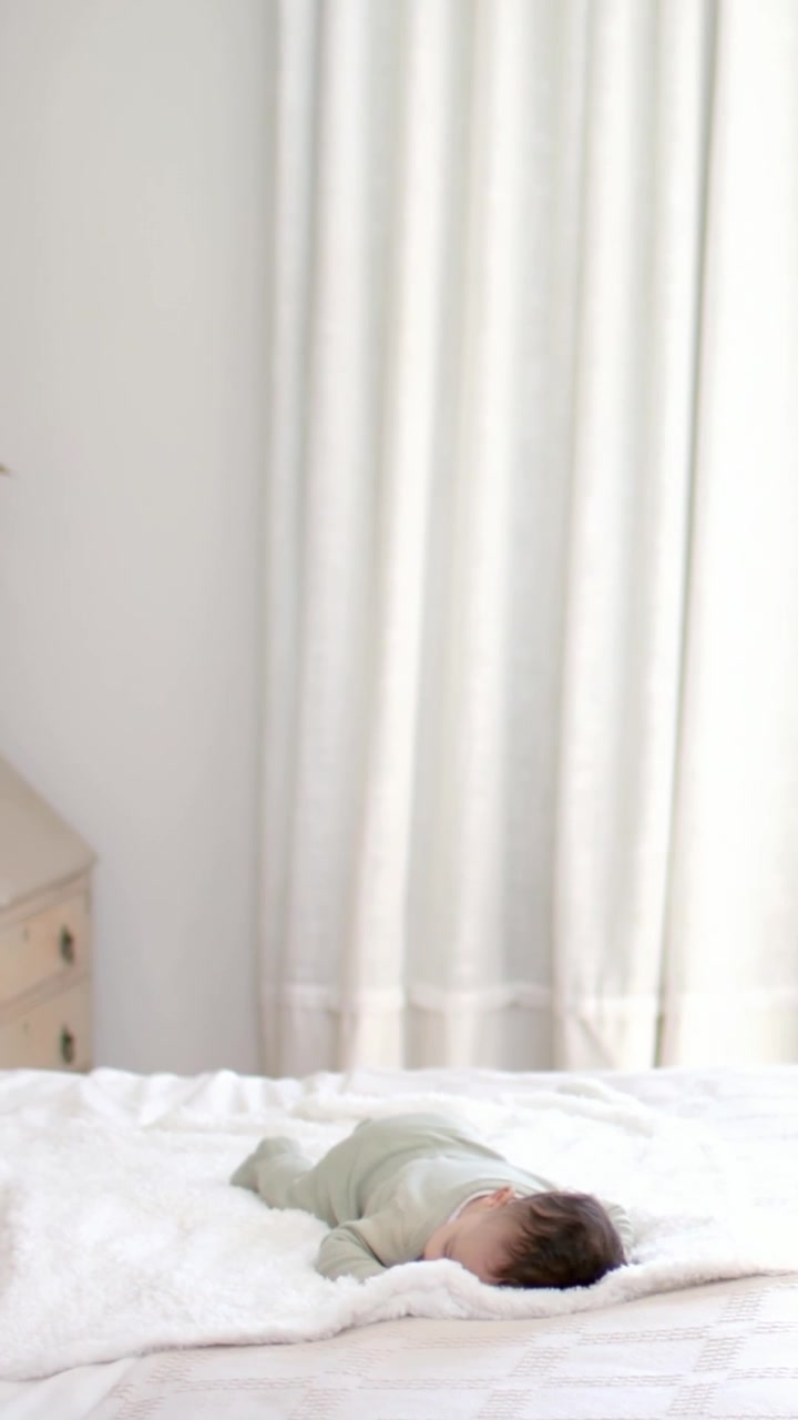 Video verticale: Genitore con maglia beige che entra e controlla un neonato in tutina che si muove leggermente su una coperta.