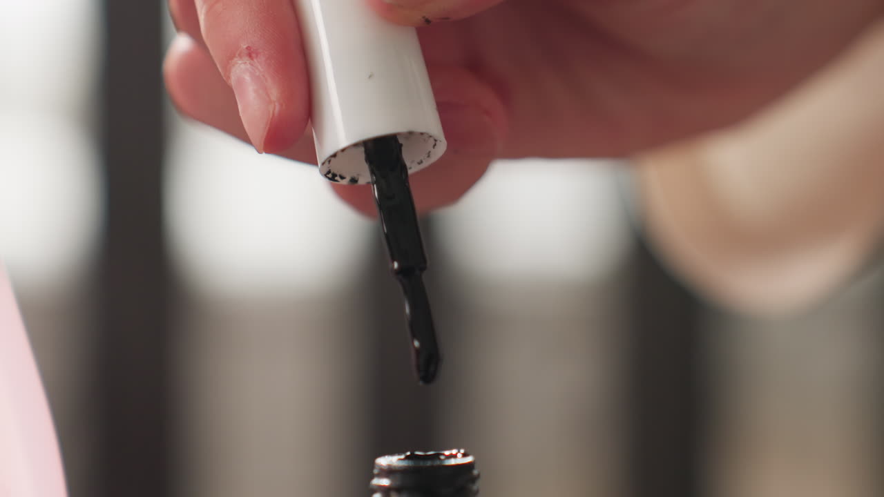 Close up teenager hands lifting white cap from black nail polish brush while drop hangs and drips back toward bottle, focus on manicure prep, beauty routine, shallow depth, indoor room table scene