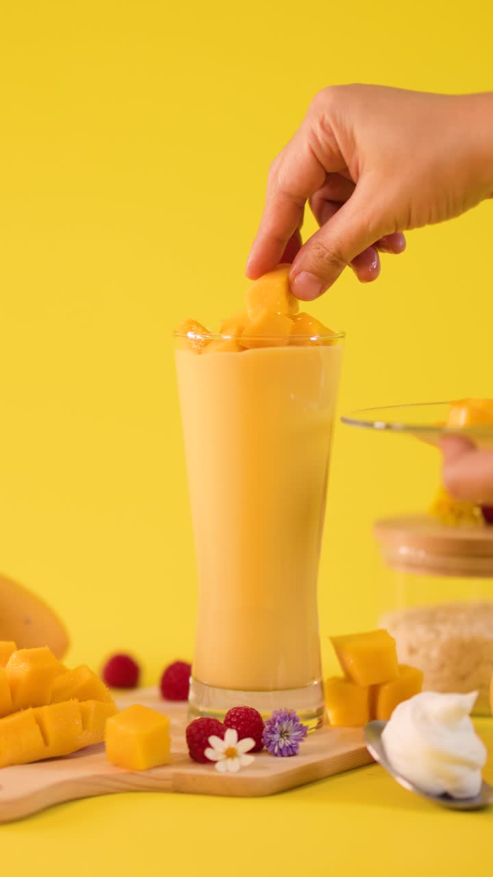 Una mano umana decora un frullato di mango alto con cubetti di frutta su uno sfondo giallo brillante in studio
