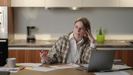 Pensive-a-woman-with-glasses-working-on-financial-documents-sitting-at-her-workplace-using-a-laptop-application-looks-focused-completing-a-task-preparing-checking-a-report-on-a-fruitful-working