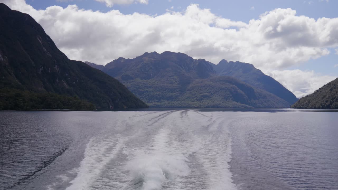 View from the back of a boat cruising through Doubtful Sound, with rippling wake, towering fjords, and calm water creating a peaceful cinematic journey