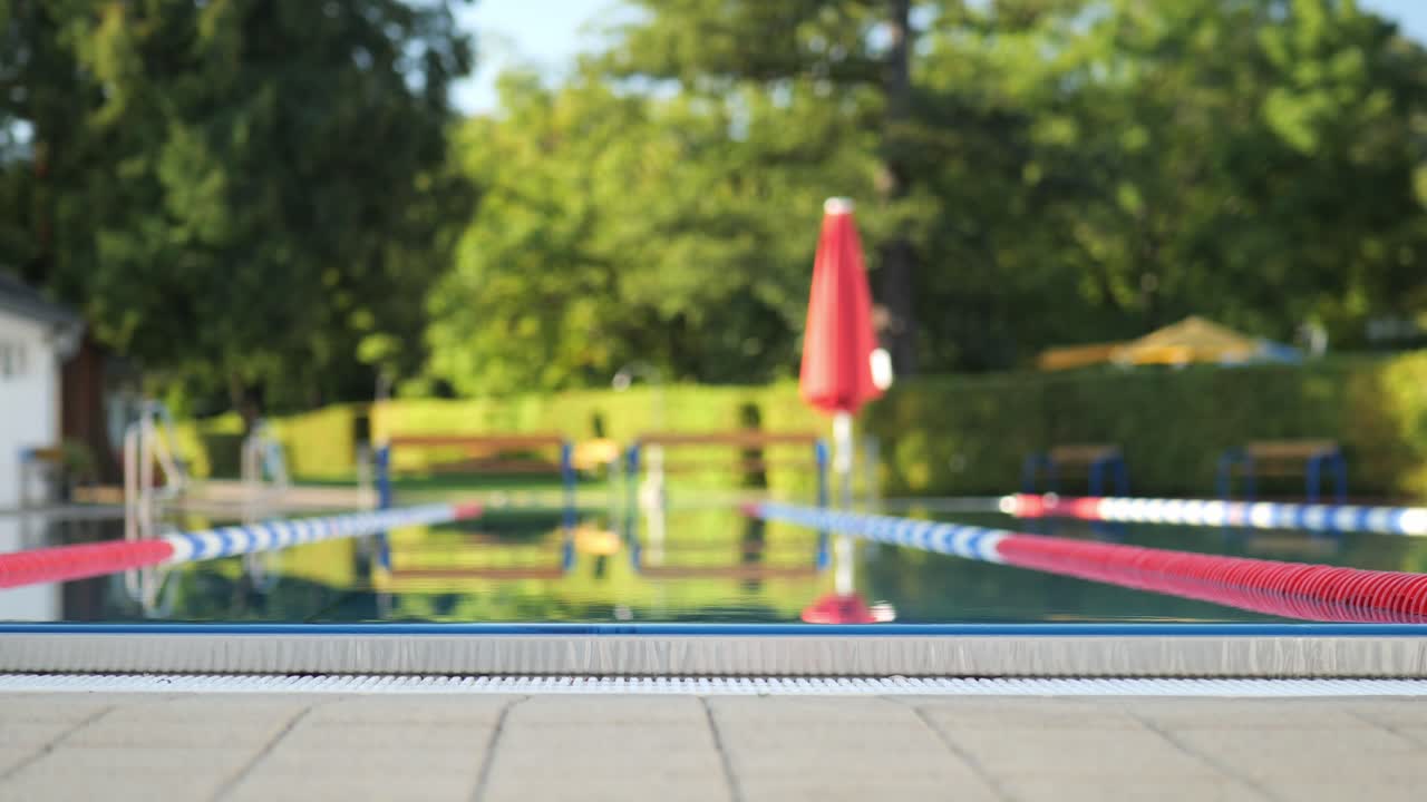 파란색 수영 트 ⁇ 크를 가진 남성 수영 선수가 수영장의 가장자리로 걸어가서 뛰어들어 수영을 시작합니다.