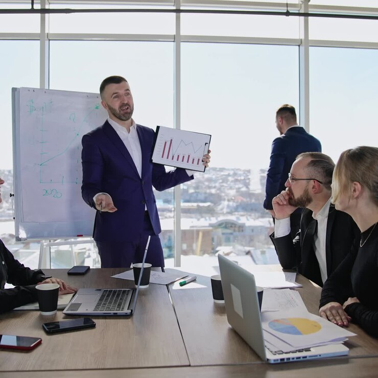 Team aziendale collaborativo riunito al tavolo per discutere le strategie. Uomo in giacca che mostra i grafici al collega. Uomo che parla al telefono sullo sfondo